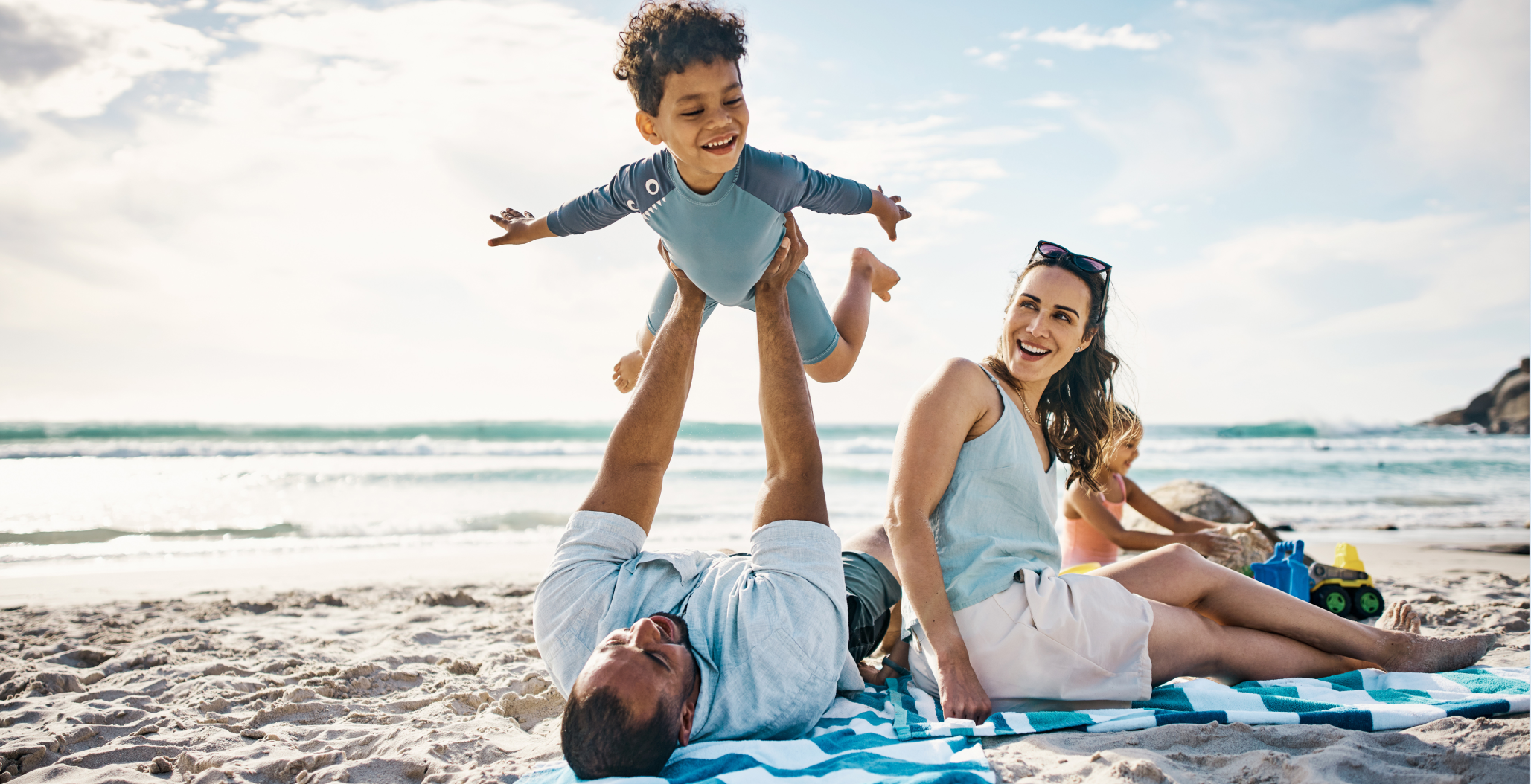 A family relaxes together on a sunny beach during Memorial Day weekend, with a father lifting a smiling child into the air while a woman and another child sit nearby on the sand.