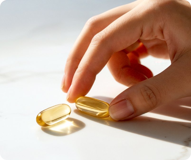 A close-up of a hand holding two golden softgel capsules against a neutral background, shown on the Metagenics OmegaGenics landing page.