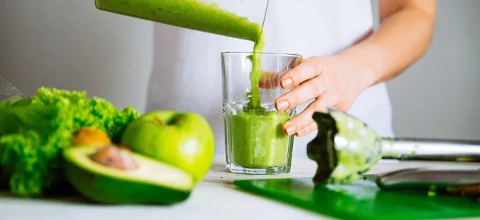 A close up of a woman pouring a green smoothie into a glass with leafy greens, an apple and an avocado in the foreground.