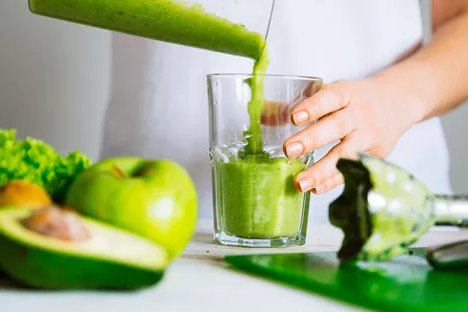 A close up of a woman pouring a green smoothie into a glass with leafy greens, an apple and an avocado in the foreground.