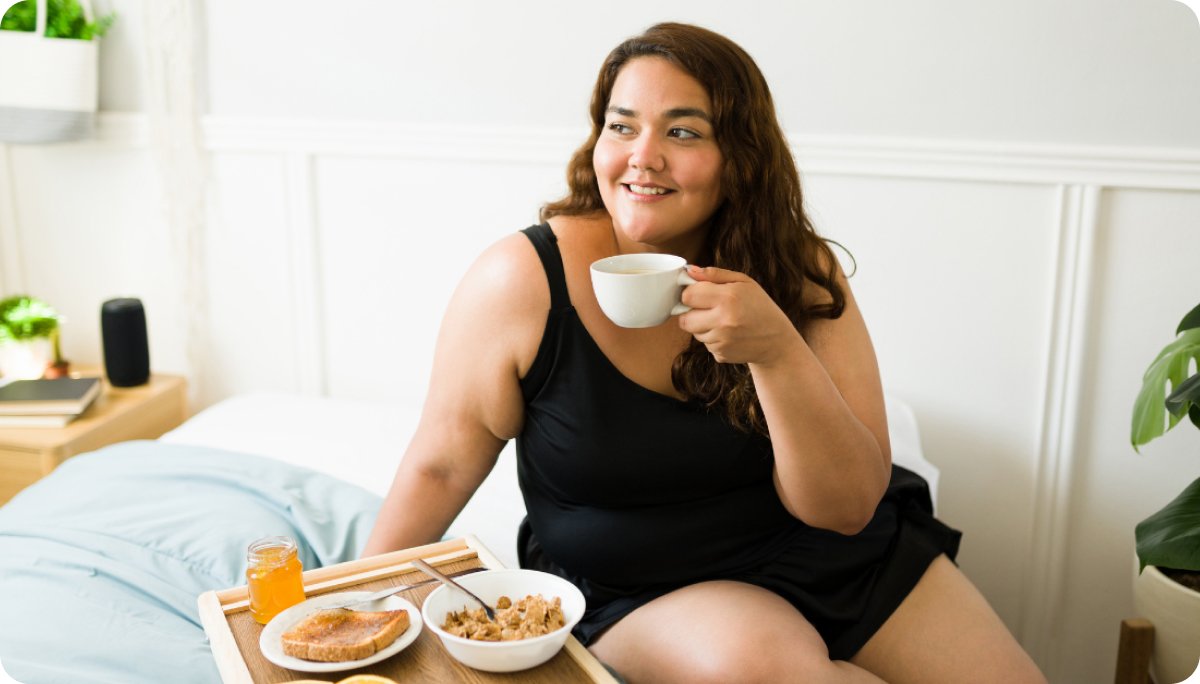 A woman sits comfortably on a bed at home holding a cup of coffee, with a breakfast tray nearby, representing the idea of building healthy daily habits and morning routines.