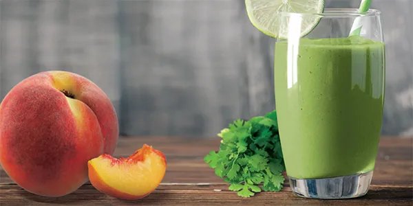 A green smoothie in a glass with a sliced peach and a bunch of cilantro on a table