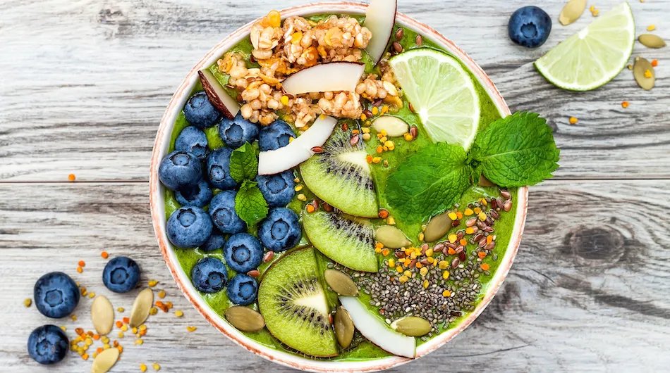 Colorful green smoothie bowl topped with blueberries, kiwi, coconut, lime, granola, seeds, and fresh mint on a rustic wooden table.