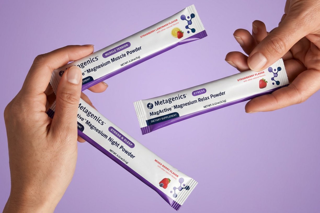 Hands holding three individually packages of Metagenics' MagActive magnesium powder supplements against a light purple background.