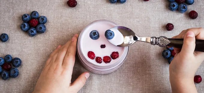 Child's hands holding a bowl of berry-topped yogurt, with a spoonful of yogurt and blueberries nearby.