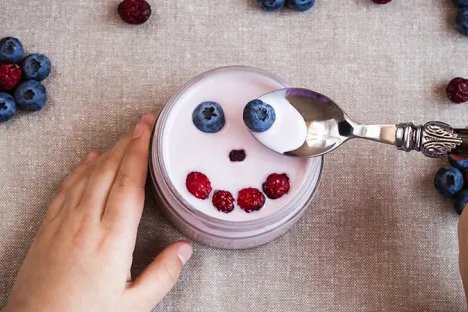 Child's hands holding a bowl of berry-topped yogurt, with a spoonful of yogurt and blueberries nearby.