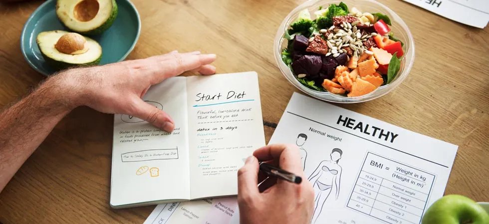 Overhead view of a person making a weightless plan in a journal with a bowl of salad and avocado on the table