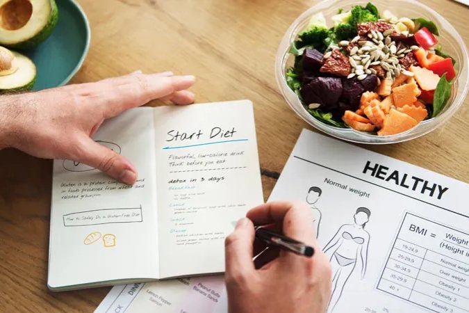 Overhead view of a person making a weightless plan in a journal with a bowl of salad and avocado on the table