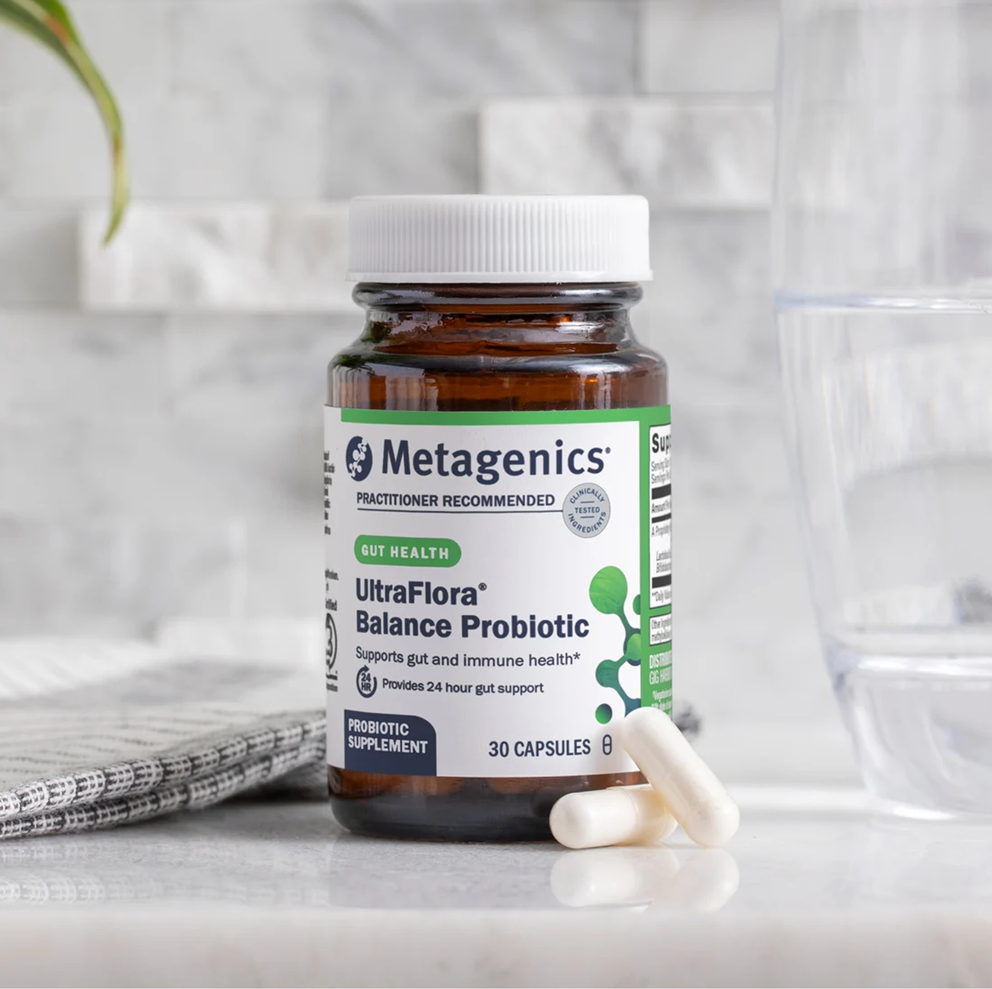 Product image of a Metagenics UltraFlora® Balance Probiotic supplement bottle on a light kitchen surface, with several white capsules placed in front of the bottle and a glass of water nearby.