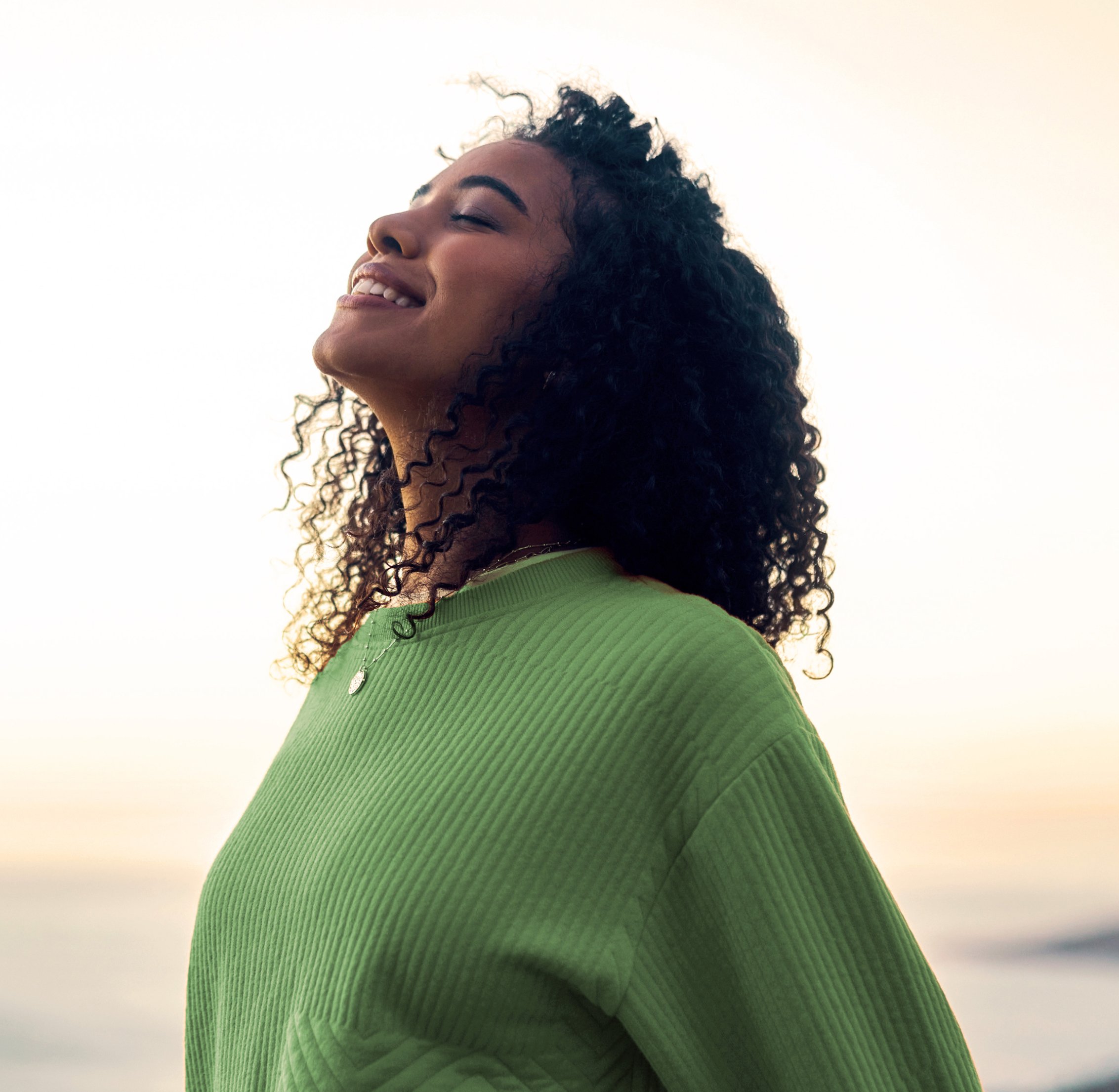 A woman wearing a green sweater stands outdoors near the water, smiling softly with her face turned upward toward the light, expressing calm, confidence, and well-being. The image reflects a sense of balance and optimism aligned with Metagenics wellness messaging, inviting viewers to reset your routine during the new year supplement sale.