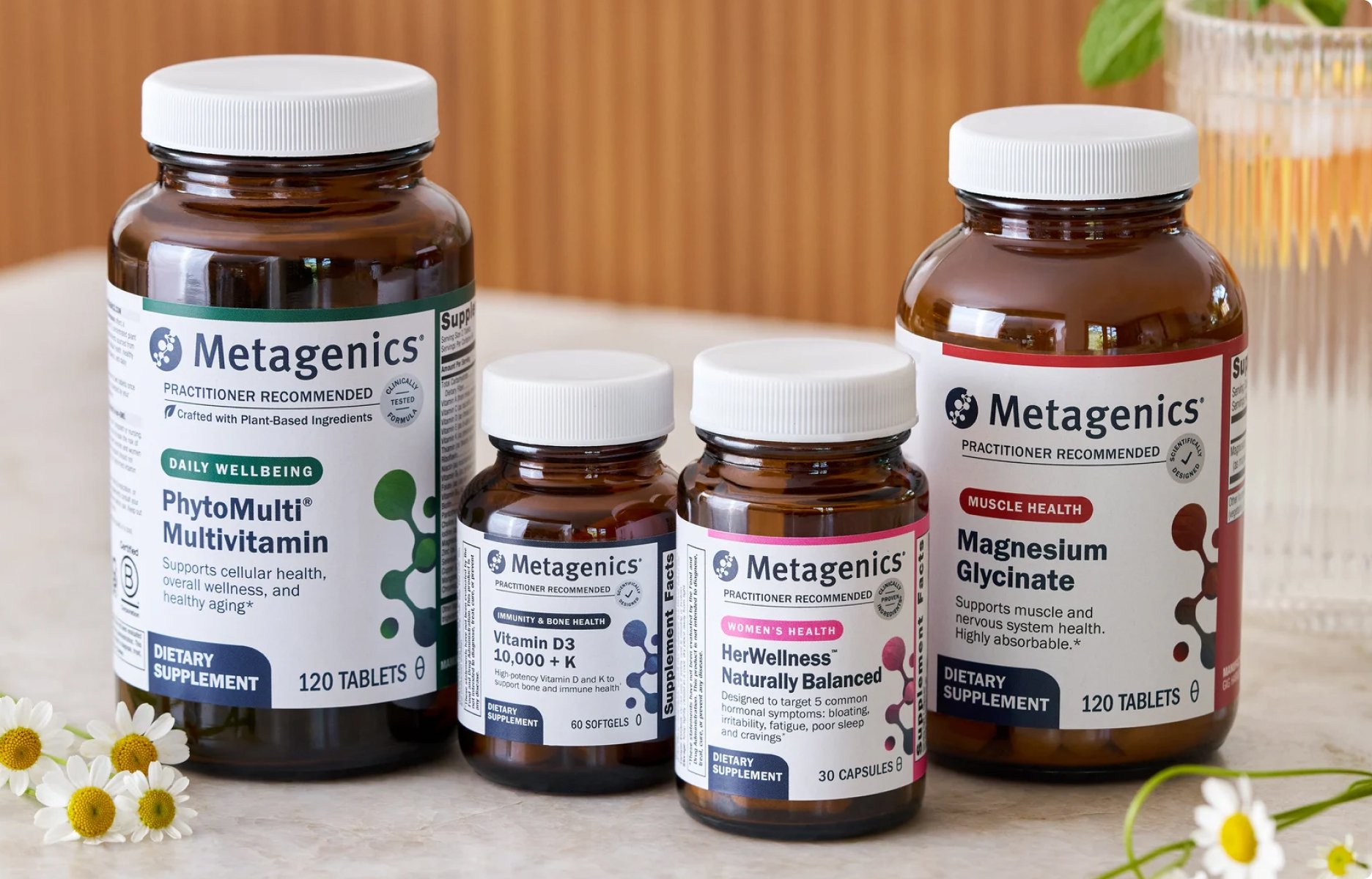 A group of Metagenics supplements displayed on a countertop, including PhytoMulti Multivitamin, Vitamin D3 10,000 + K, HerWellness Naturally Balanced, and Magnesium Glycinate. The amber bottles with white caps are arranged together with small flowers nearby, creating a clean wellness aesthetic. This image represents FSA/HSA Eligible Vitamins & Supplements, highlighting that customers may be able to use HSA or FSA funds to purchase Metagenics products.