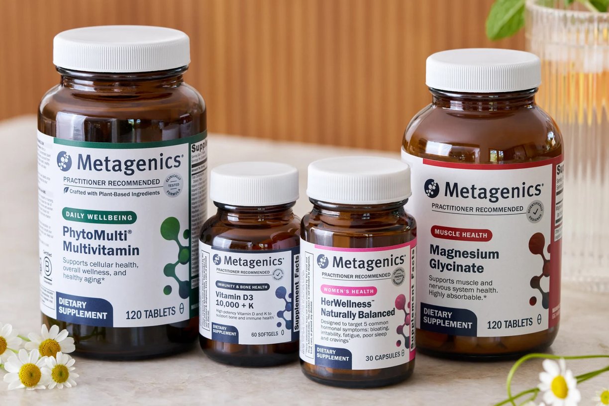 A group of Metagenics supplements displayed on a countertop, including PhytoMulti Multivitamin, Vitamin D3 10,000 + K, HerWellness Naturally Balanced, and Magnesium Glycinate. The amber bottles with white caps are arranged together with small flowers nearby, creating a clean wellness aesthetic. This image represents FSA/HSA Eligible Vitamins & Supplements, highlighting that customers may be able to use HSA or FSA funds to purchase Metagenics products.