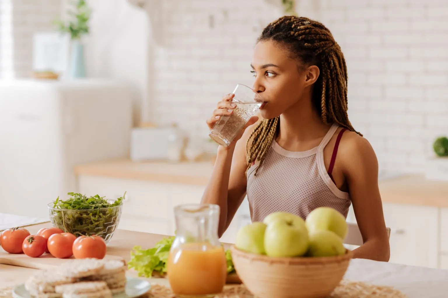 Woman drinking water at a kitchen counter with fresh fruits and vegetables.