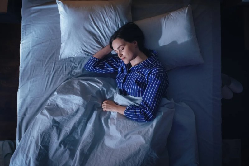 A woman in blue striped pajamas lies asleep in bed under a light-colored blanket, viewed from above in dim blue-toned lighting.