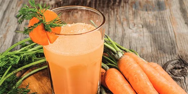 An orange smoothie in a glass on a table next to a bunch of fresh carrots