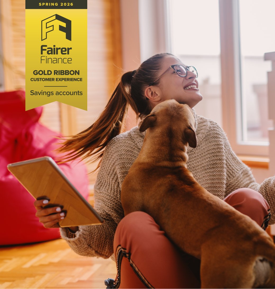 Woman sitting on the floor and playing with dog. In hand tablet. Living room interior. Fairer Finance Gold Ribbon in the top left corner of the image for savings customer experience.
