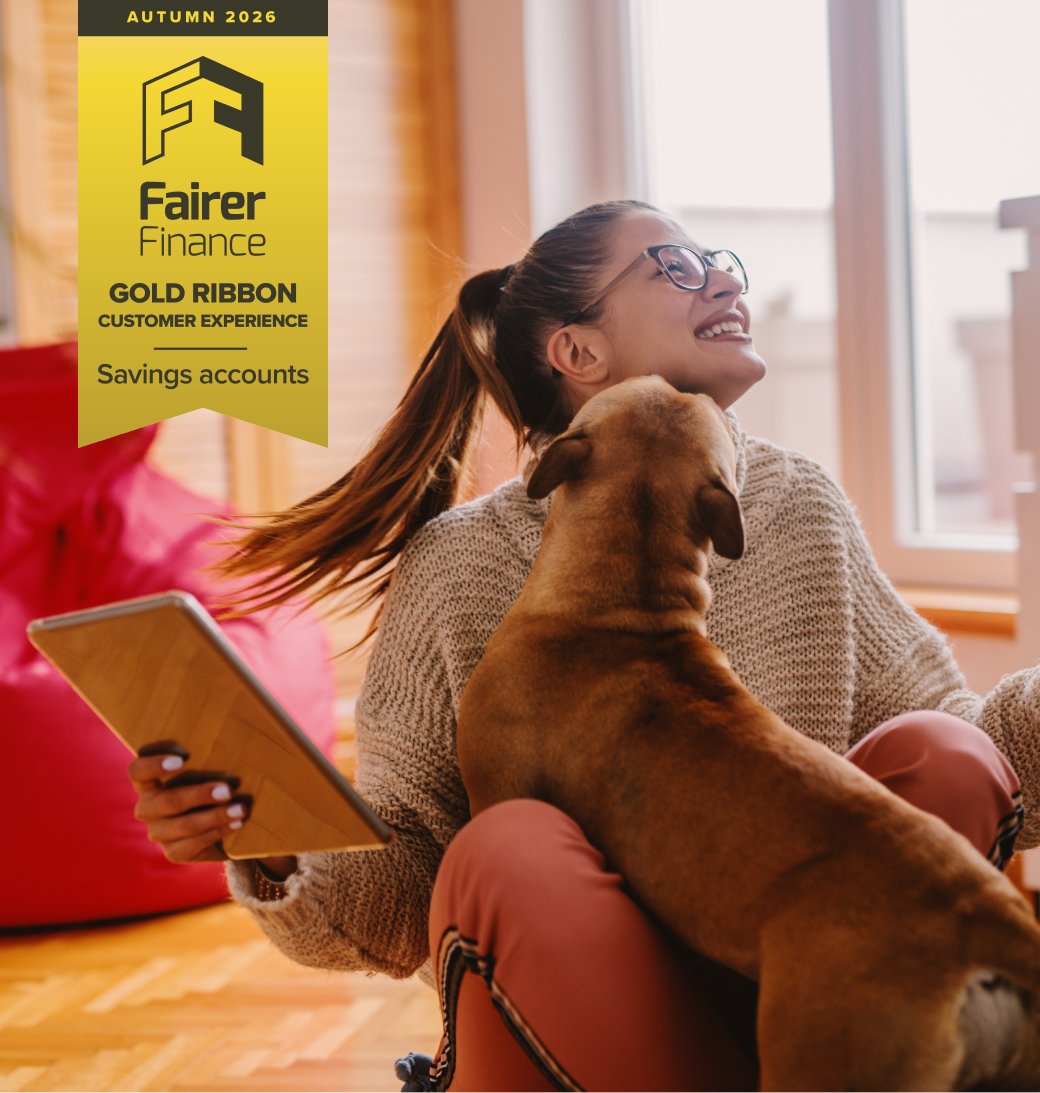 Woman sitting on the floor and playing with dog. In hand tablet. Living room interior. Fairer Finance Gold Ribbon in the top left corner of the image for savings customer experience.