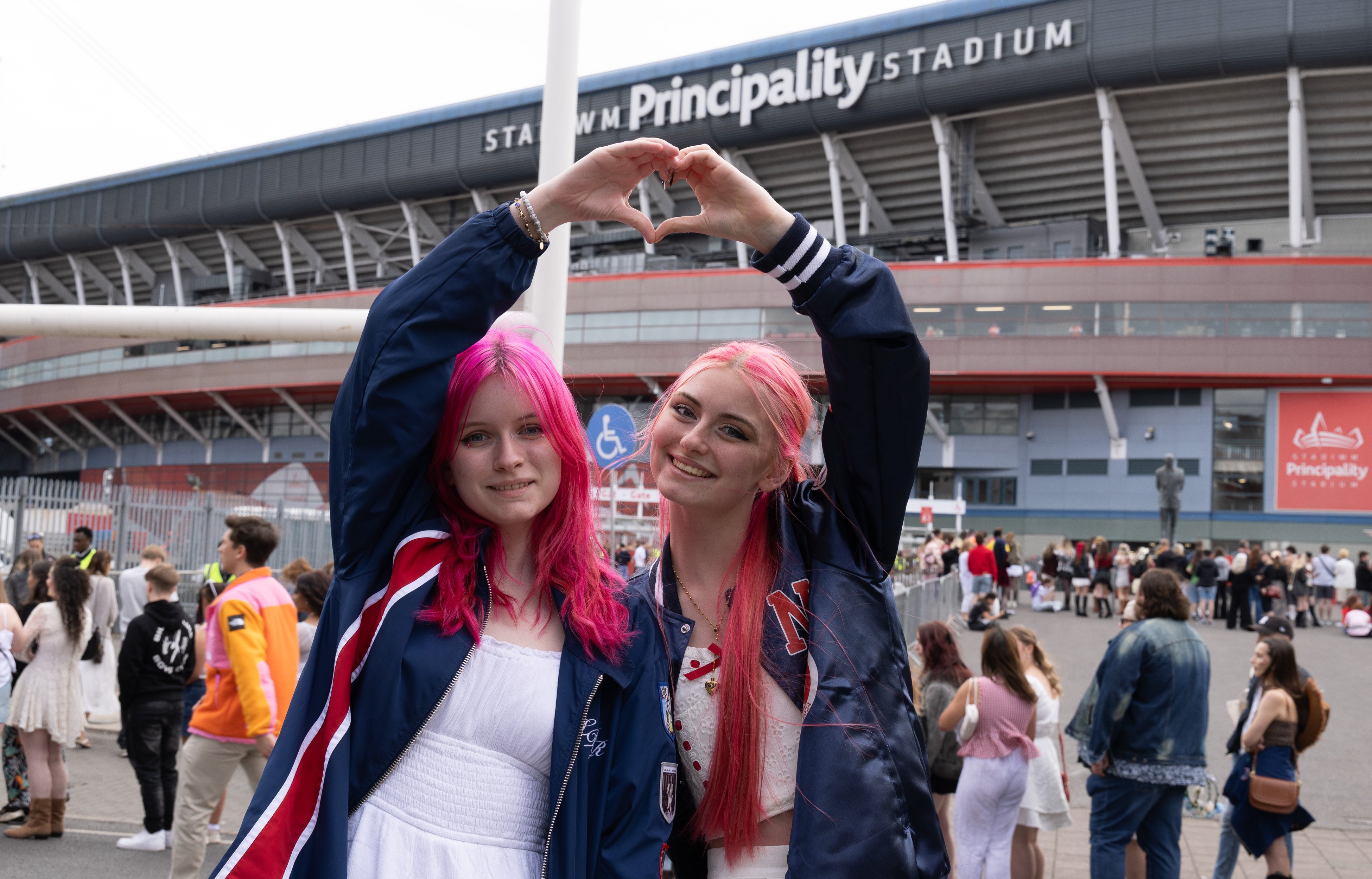 Two young people with bright pink hair stand outside Principality Stadium, smiling and forming a heart shape with their arms. They are wearing navy jackets over light outfits, and a crowd of people is gathered in the background near the stadium entrance.