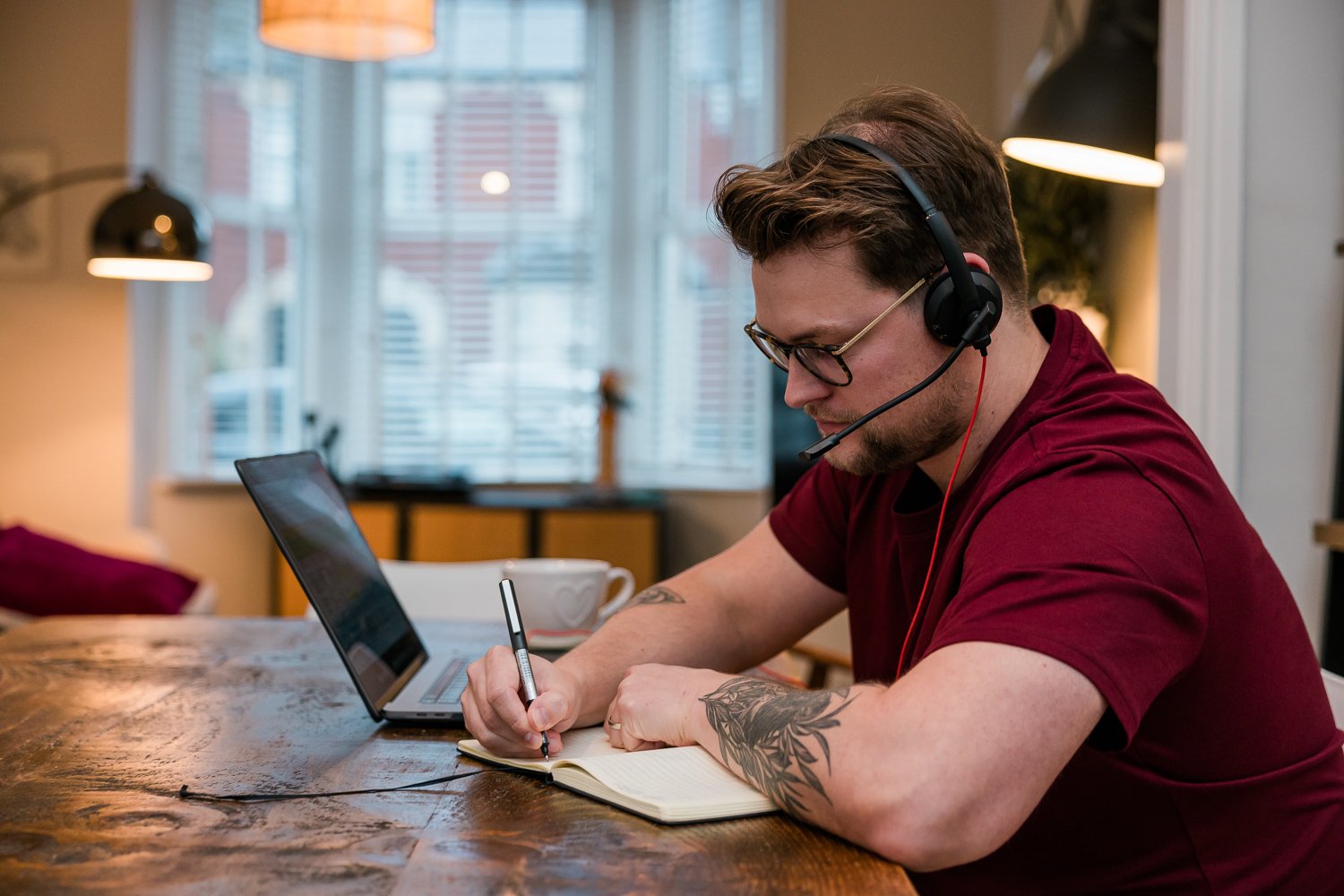Man sits at desk with laptop, pen and notepad.  He wears headphones.