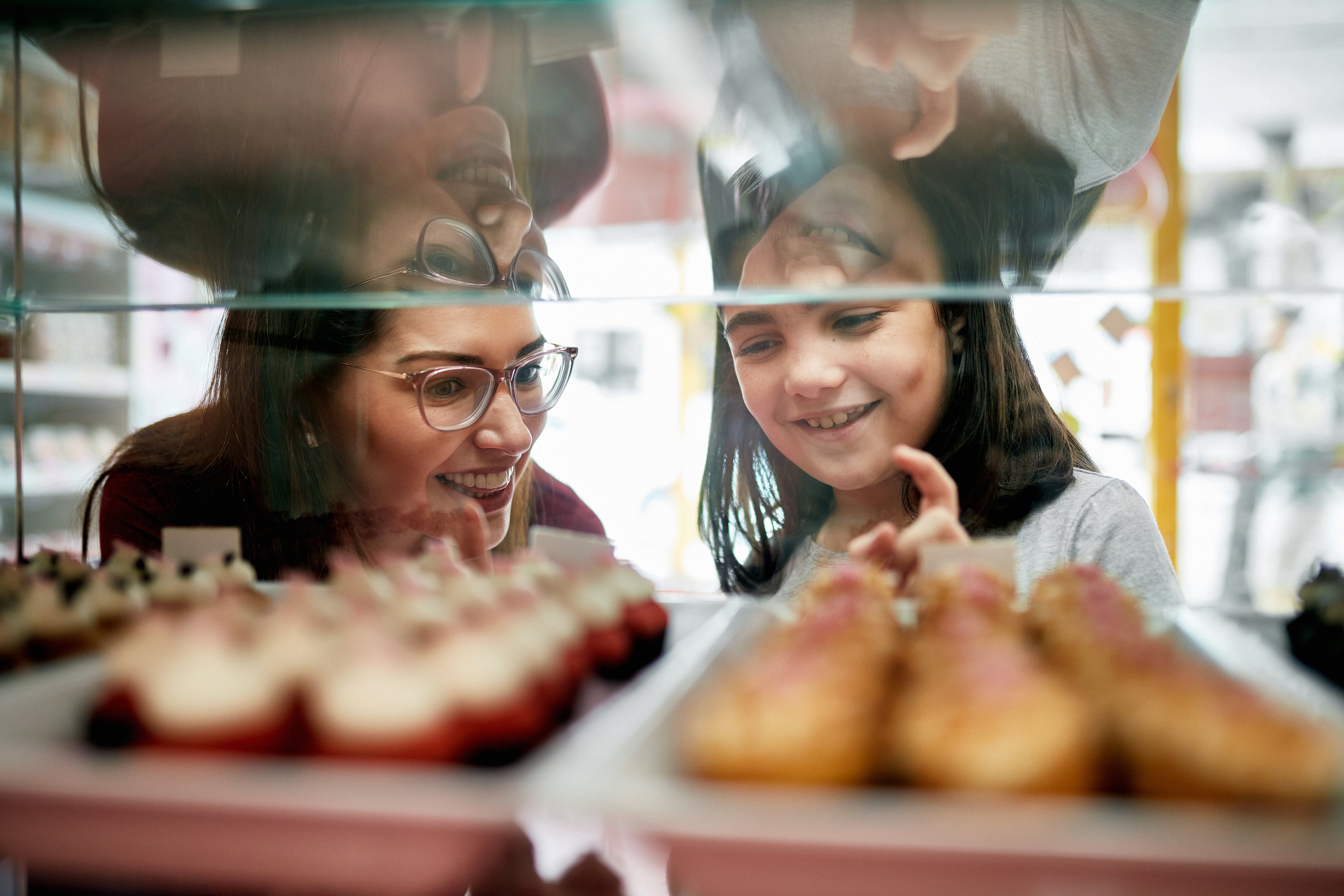 Focus on background of mother and 9 year old daughter smiling as they pick gourmet cupcakes from bakery display case.