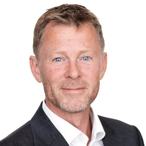 A person with short hair and a beard wearing a dark suit jacket and white shirt.
