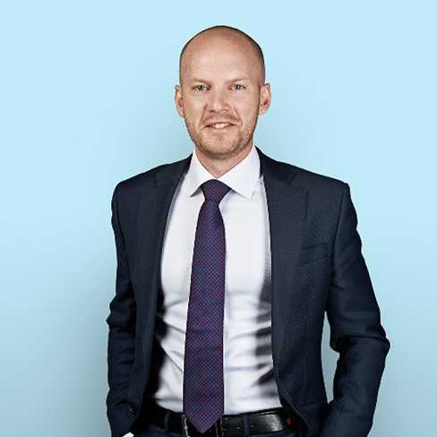 A person wearing a dark suit and tie standing against a light blue background.