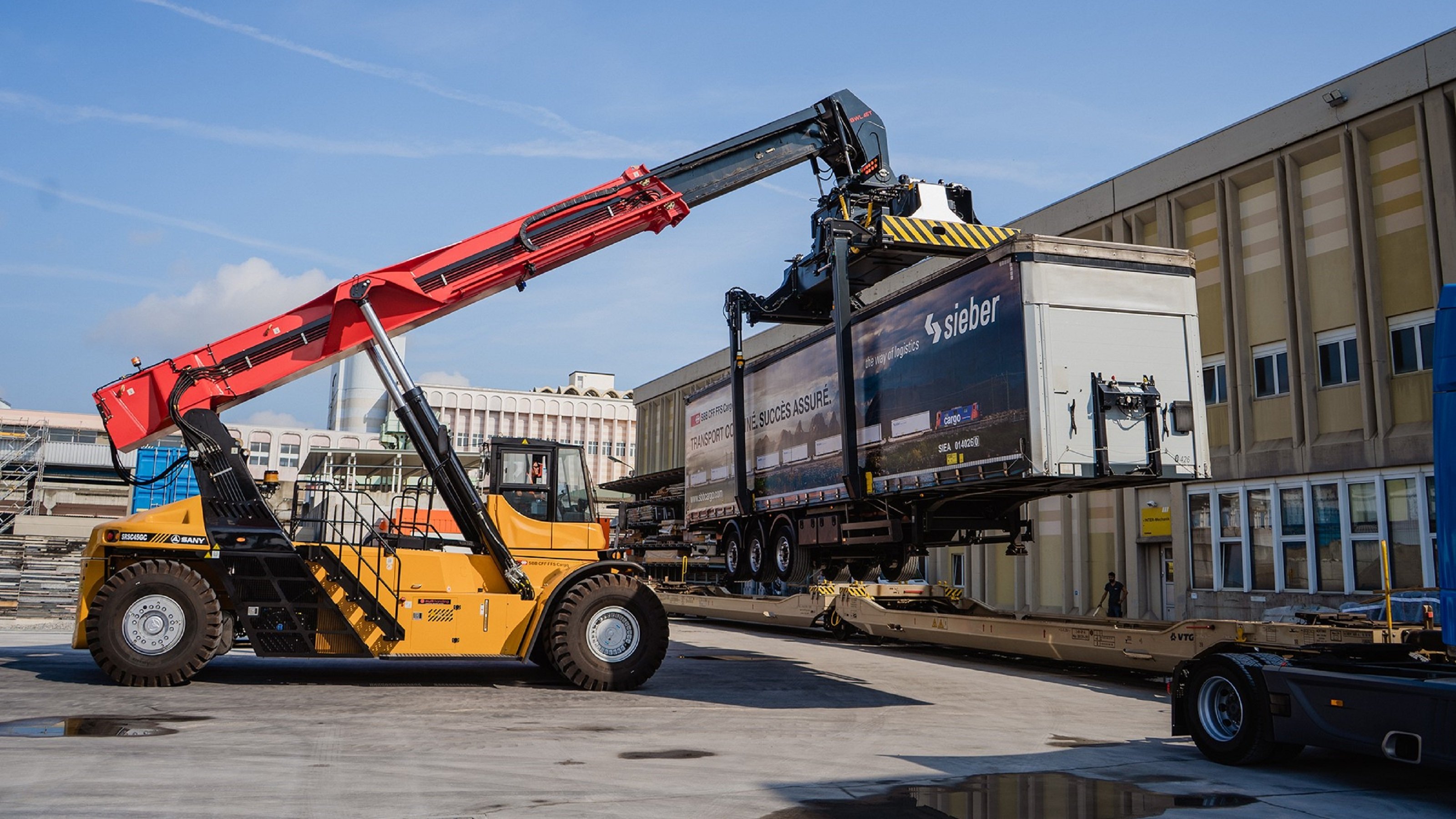 Alternativtext für Sehbehinderte (max. 2-3 Sätze): Ein grosser gelber Mobilkran mit einem ausgestreckten roten Arm hebt einen Sattelauflieger mit der Aufschrift “Sieber” in die Luft, der auf einem Anhänger steht. Der Kran arbeitet auf einem gepflasterten Aussenbereich, umgeben von Gebäuden und Strukturen unter einem klaren blauen Himmel.