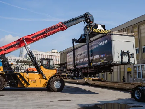 Alternativtext für Sehbehinderte (max. 2-3 Sätze): Ein grosser gelber Mobilkran mit einem ausgestreckten roten Arm hebt einen Sattelauflieger mit der Aufschrift “Sieber” in die Luft, der auf einem Anhänger steht. Der Kran arbeitet auf einem gepflasterten Aussenbereich, umgeben von Gebäuden und Strukturen unter einem klaren blauen Himmel.