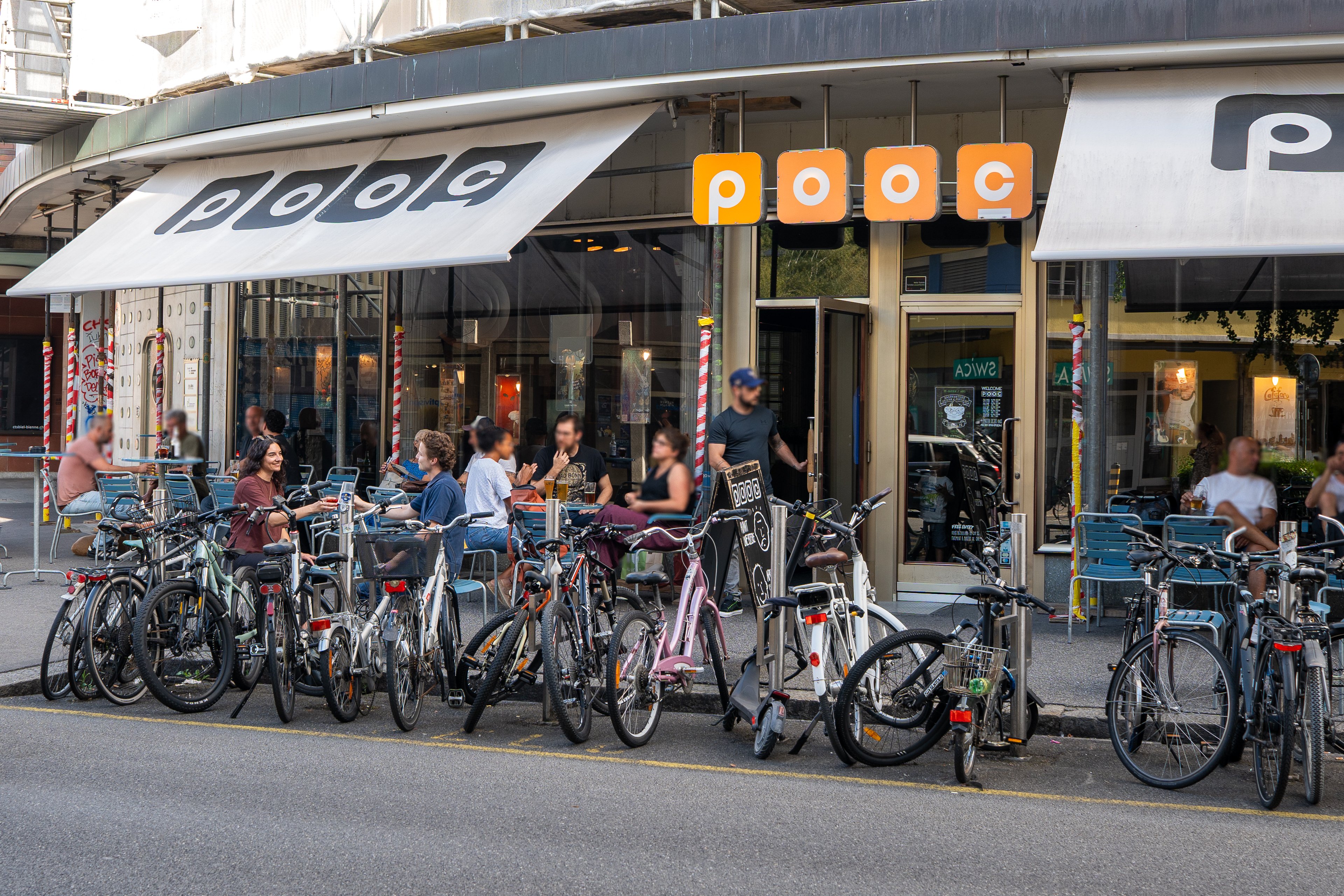 Su una strada, numerose biciclette sono allineate una accanto all’altra. Dietro di esse, diverse persone sono sedute a piccoli tavoli davanti a un bar con un’ampia vetrata.