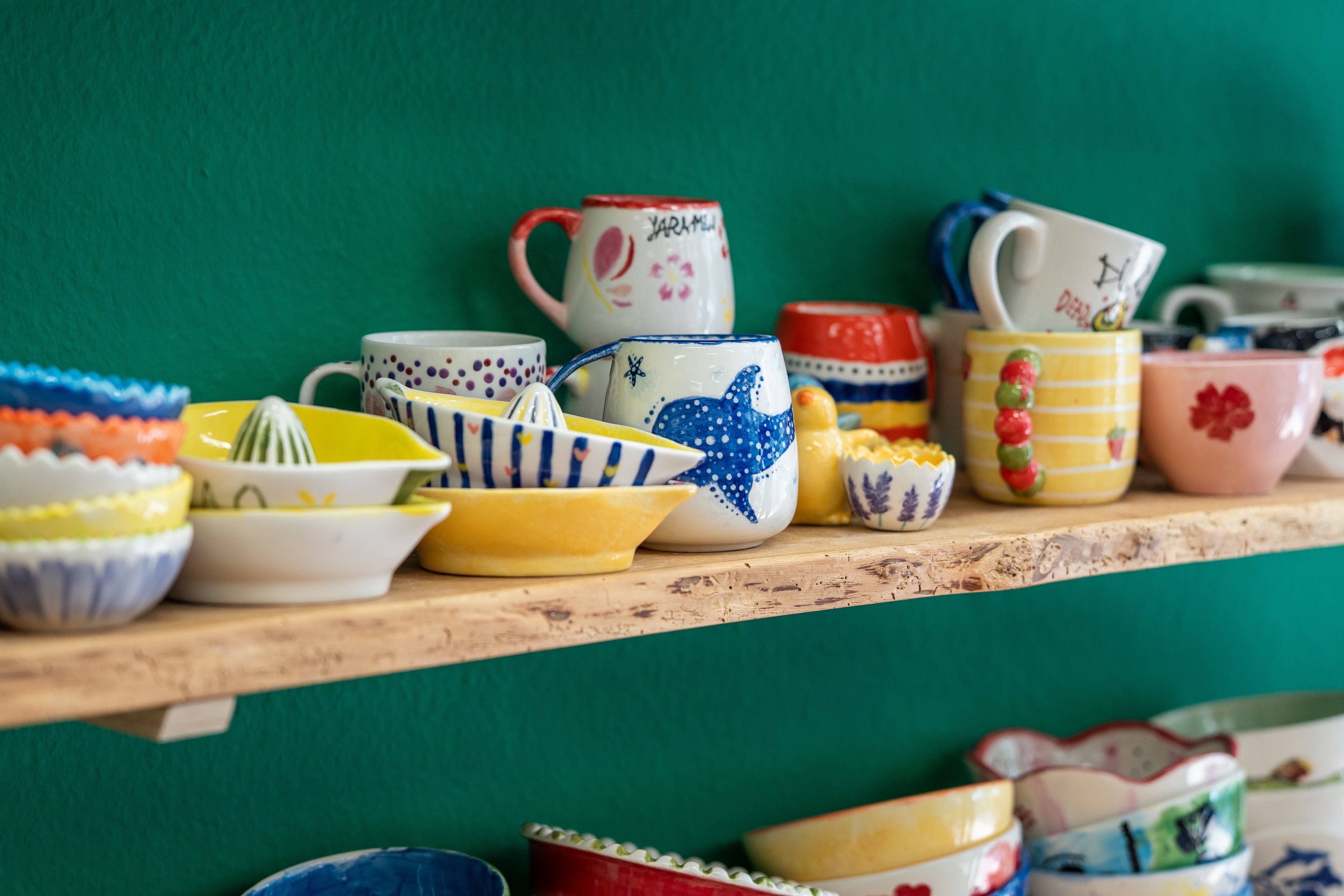 Su uno scaffale si trovano numerosi oggetti in ceramica dipinti con colori vivaci e decorazioni personalizzate. Sullo sfondo si vede una parete verde brillante.