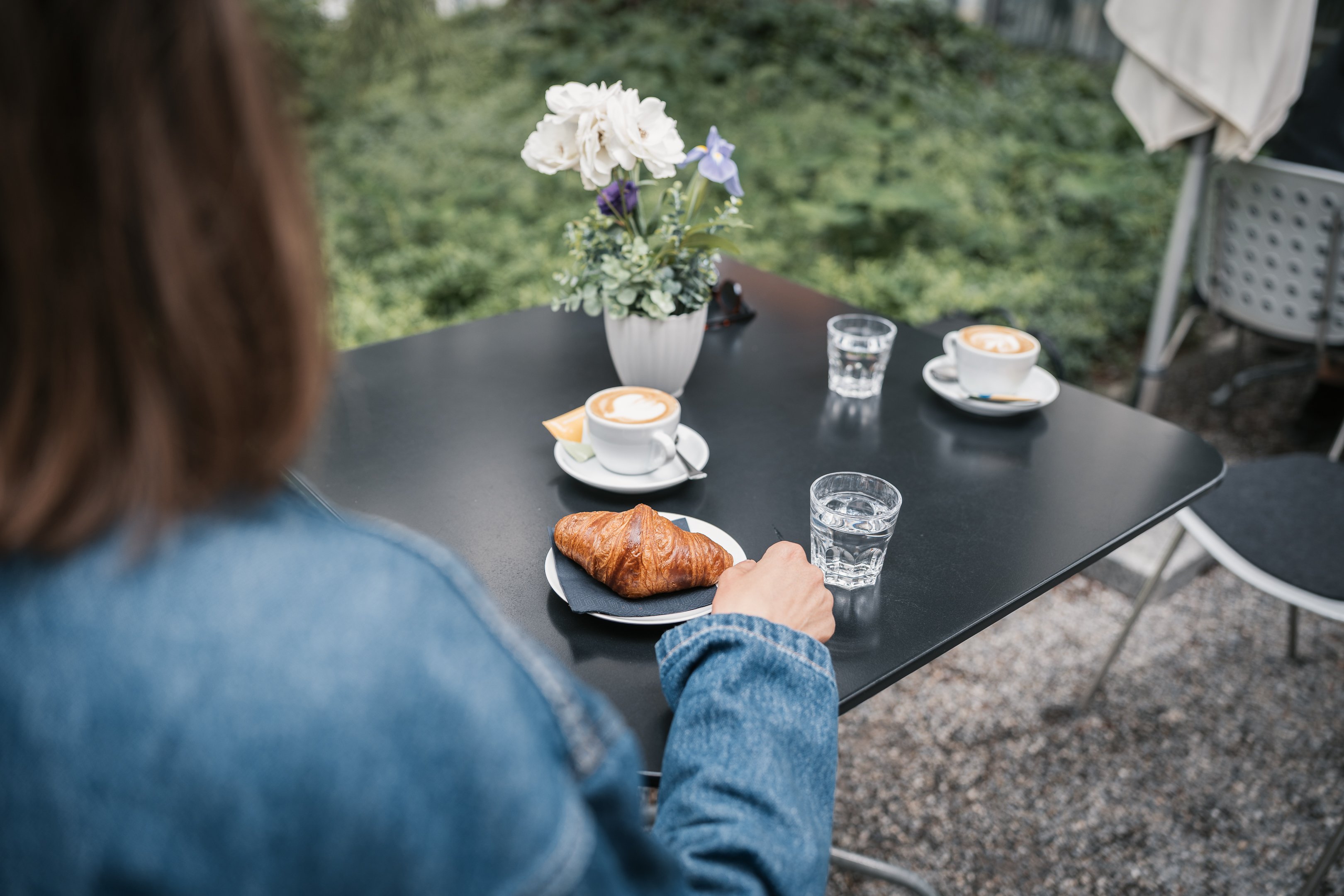 Una donna seduta a un tavolo in giardino gusta un cappuccino e un cornetto al burro.