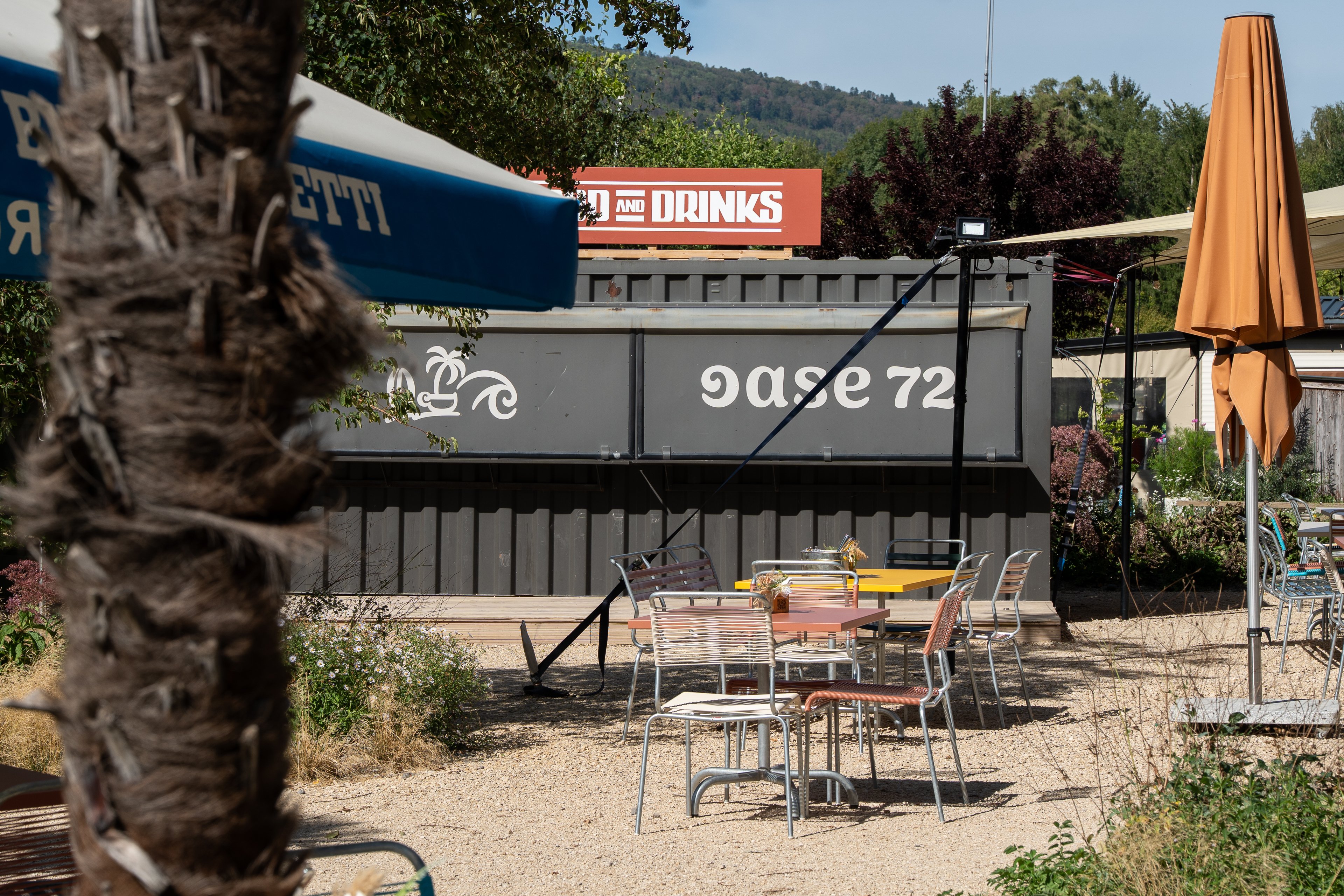 Tavoli vuoti su uno spiazzo di ghiaia dell’Oase 72 sul lago di Bienne. Sullo sfondo si trova un container con la scritta «Oase 72»; a sinistra si vede il tronco di una palma.