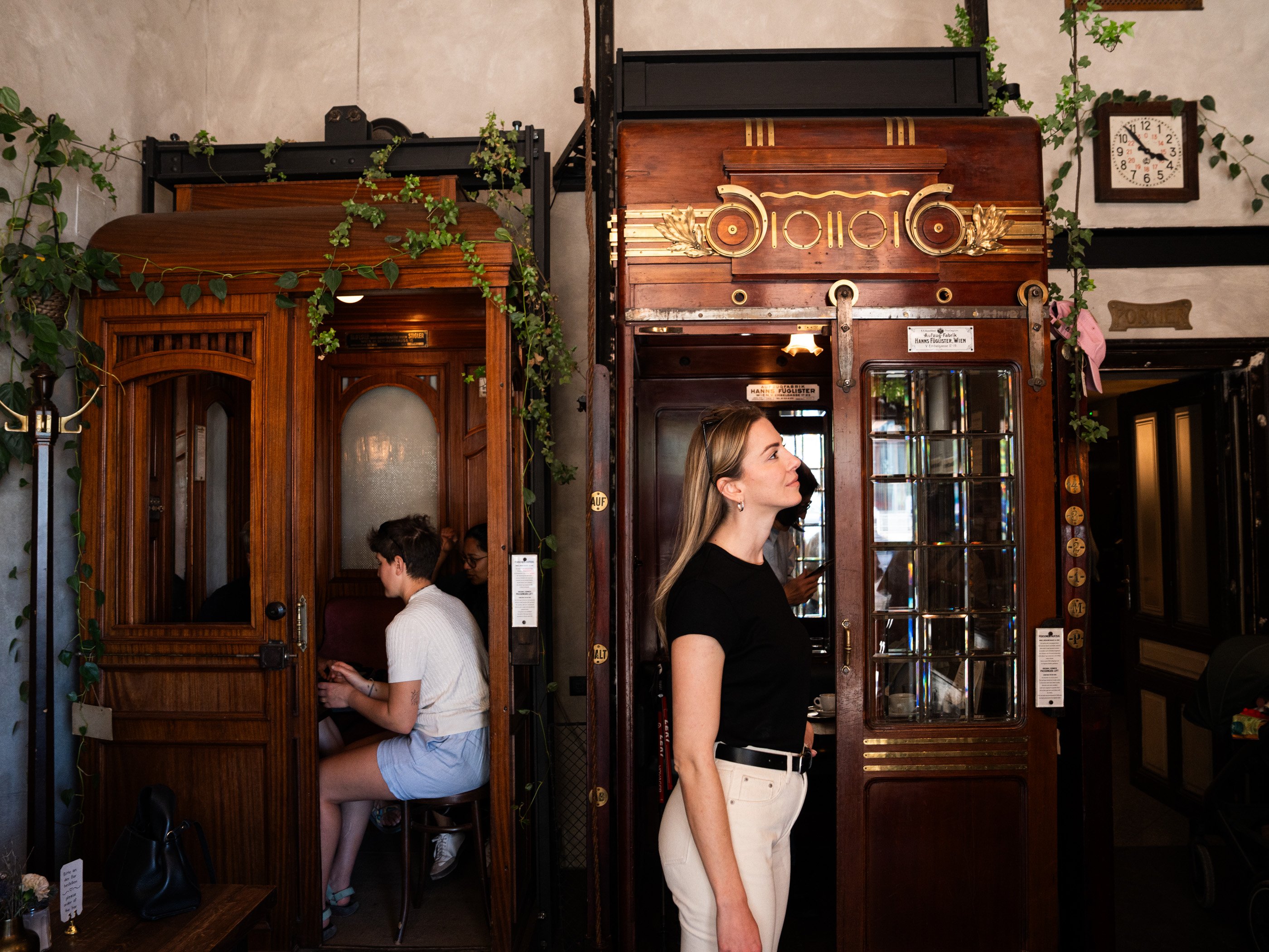 Una donna è in piedi davanti a due storiche cabine ascensore nell’Aufzug Café di Vienna, dove alcuni clienti stanno sorseggiando un caffè.