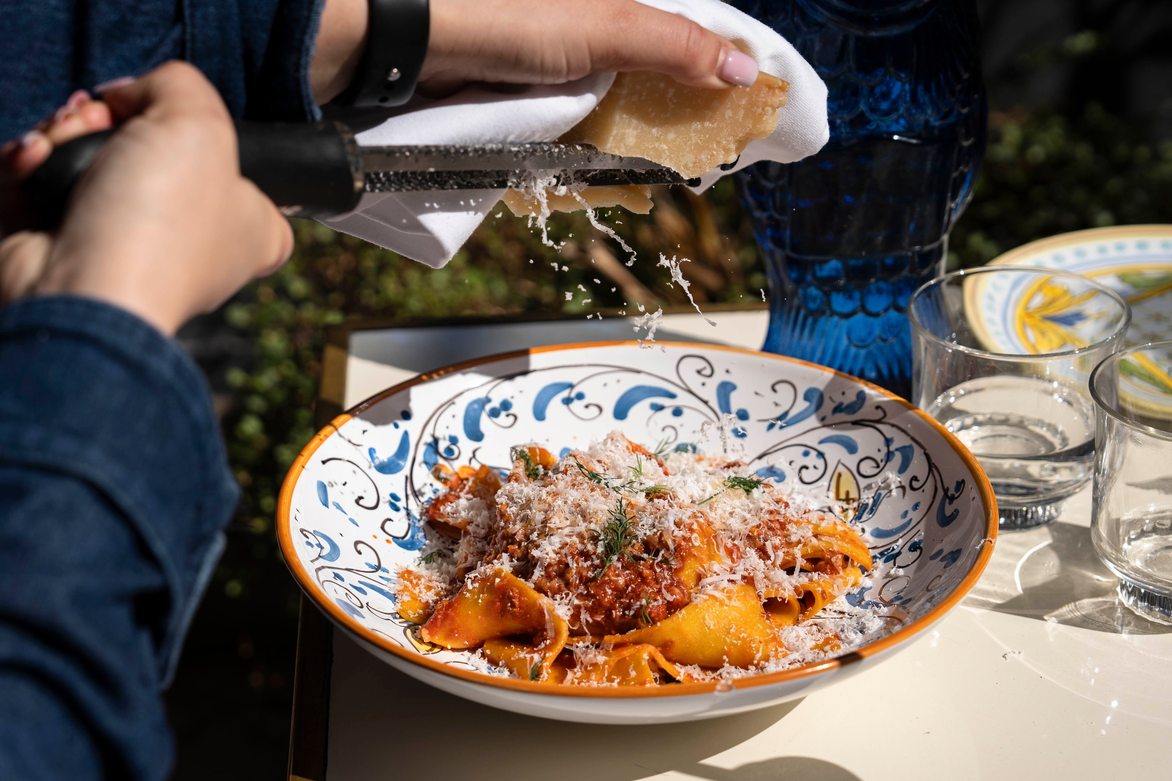 Mains râpant du parmesan au-dessus d’un bol de pâtes italiennes et motif de vaisselle rehaussant le charme du plat.