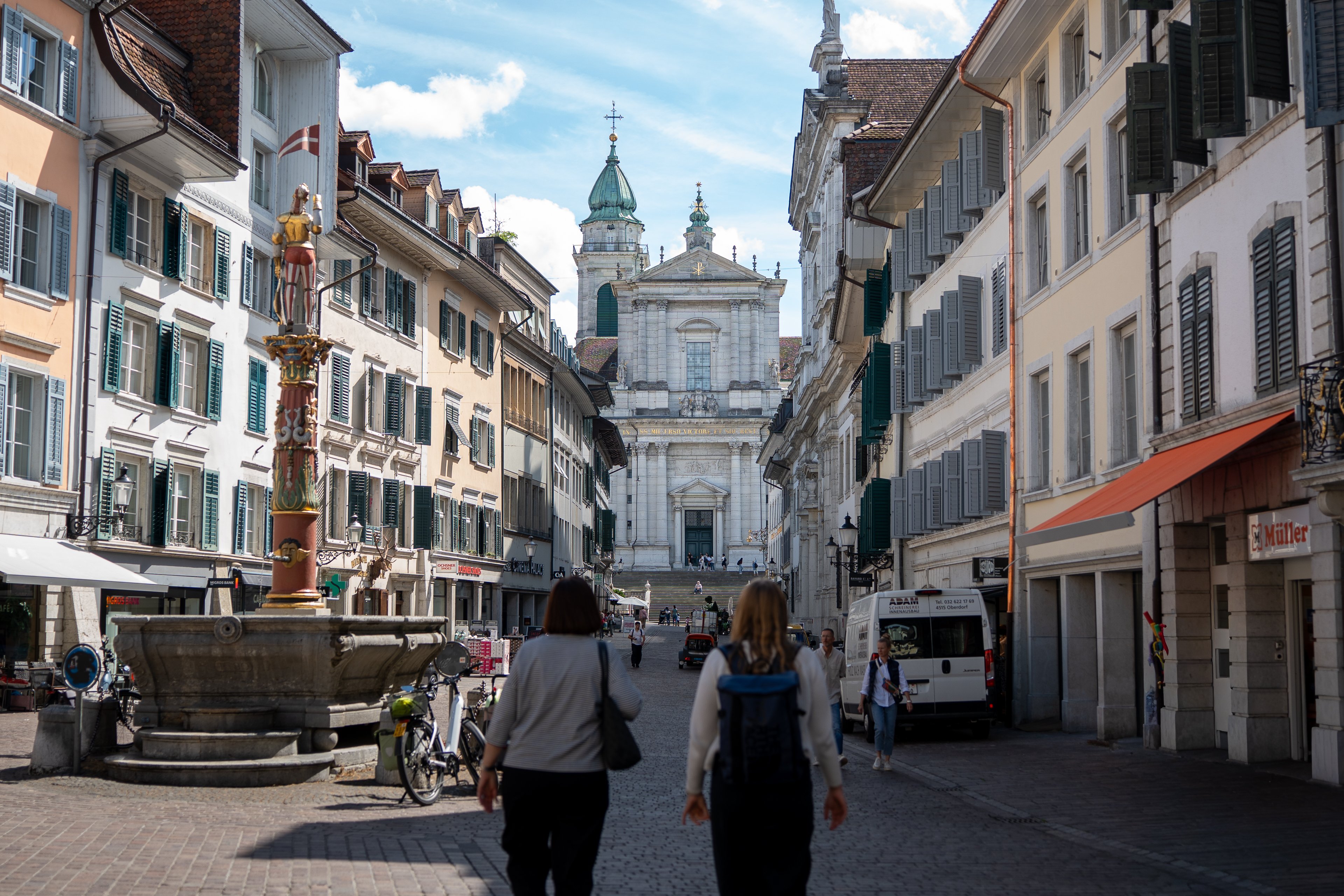 Zwei Menschen in einer breiten Altstadtgasse, in der sich ein Brunnen befindet. Am Ende der Gasse ist die Fassade einer grossen Kirche und ein Turm zu sehen.