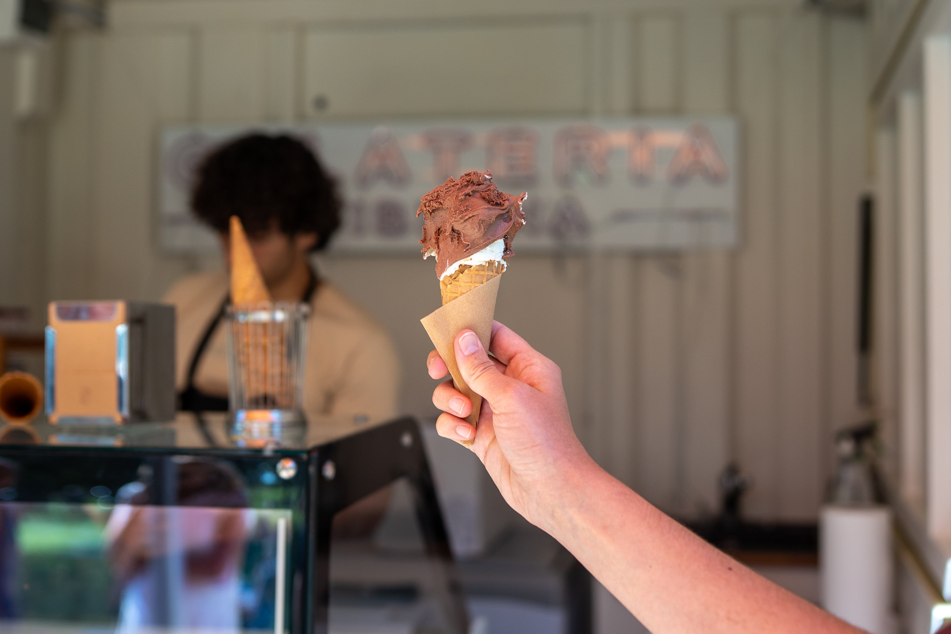 Una persona tiene in mano un gelato. Sullo sfondo si vede l’immagine sfocata del chiosco dei gelati dell’Oase 72.