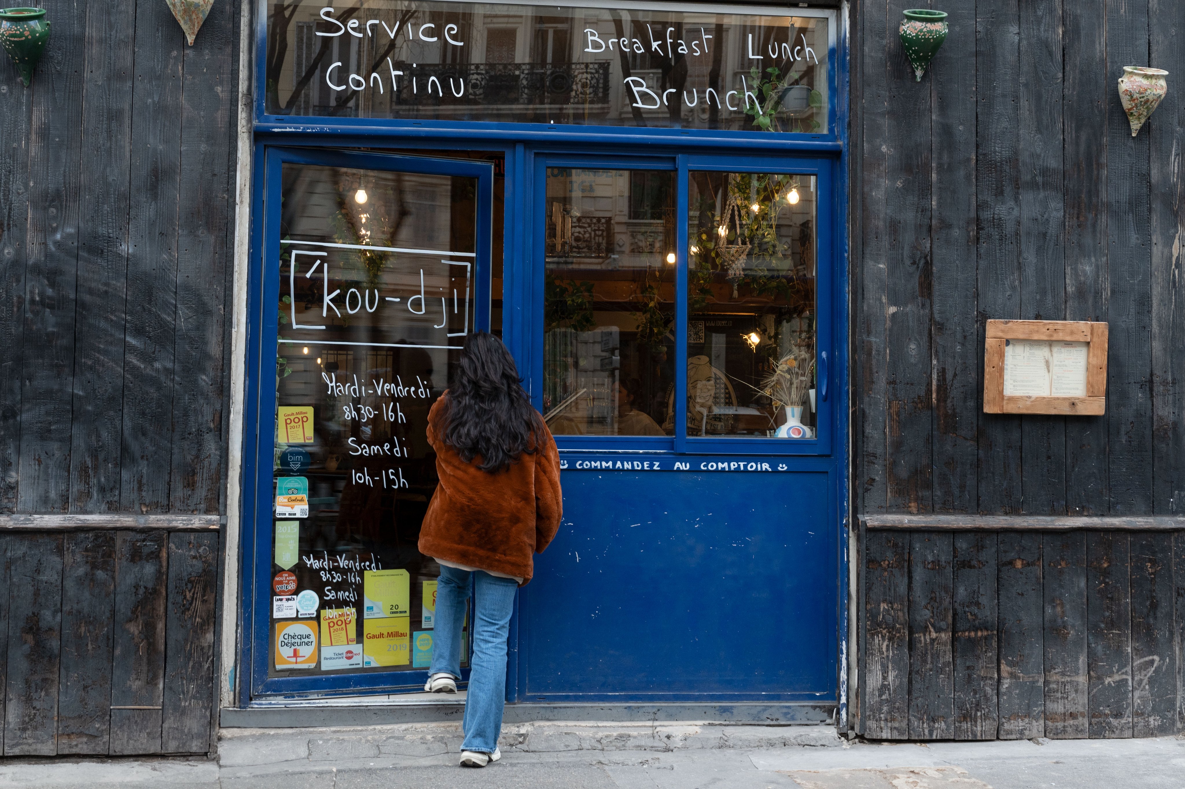 Une personne devant le café Coogee, à Marseille, regardant l’ardoise avec les horaires d’ouverture en continu ainsi que les offres pour le petit-déjeuner, le dîner et le brunch.