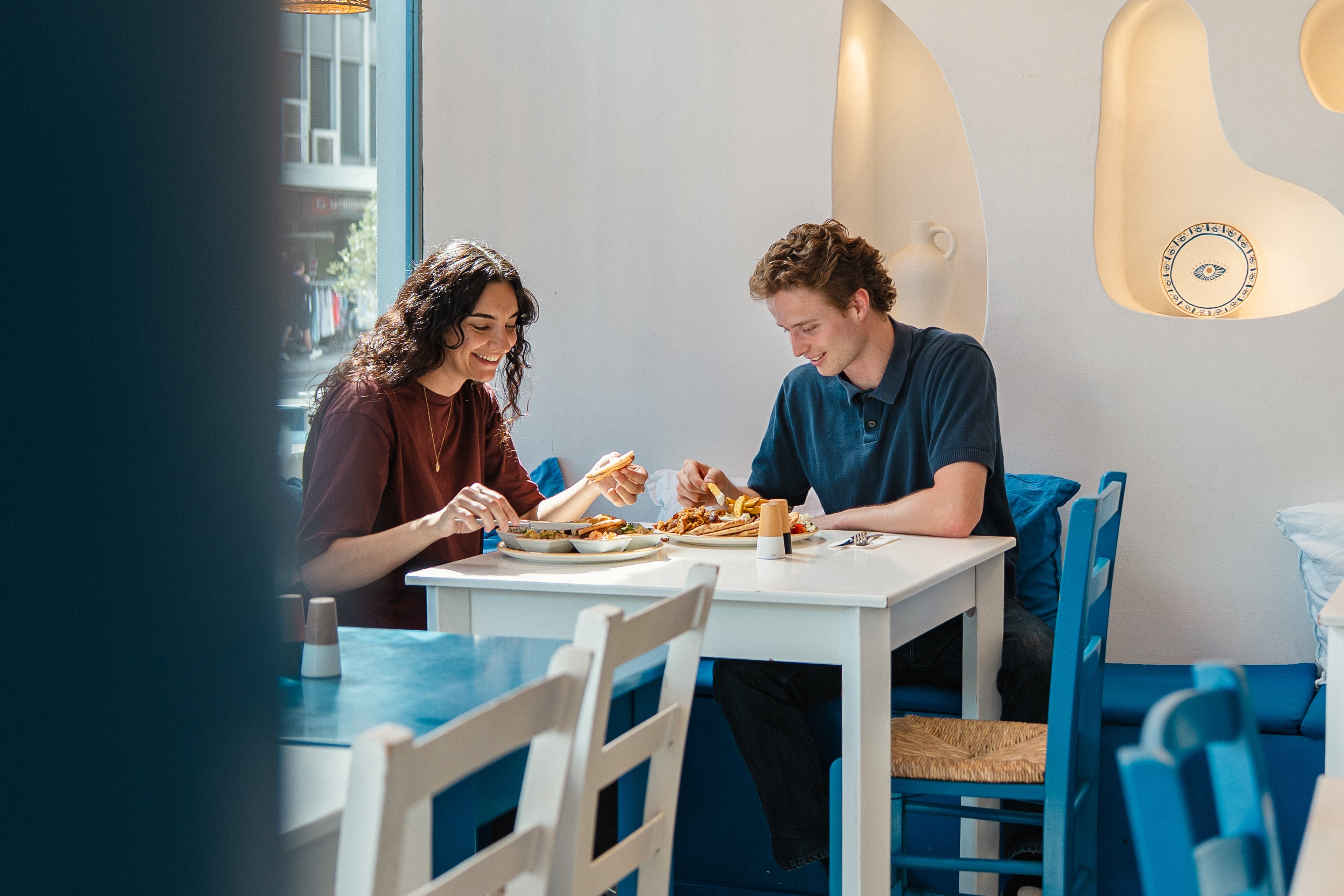 Due persone sono sedute a un tavolo del ristorante Greeka. Davanti a loro ci sono piatti con specialità greche. I tavoli e le sedie del locale sono nei toni del bianco e del blu.