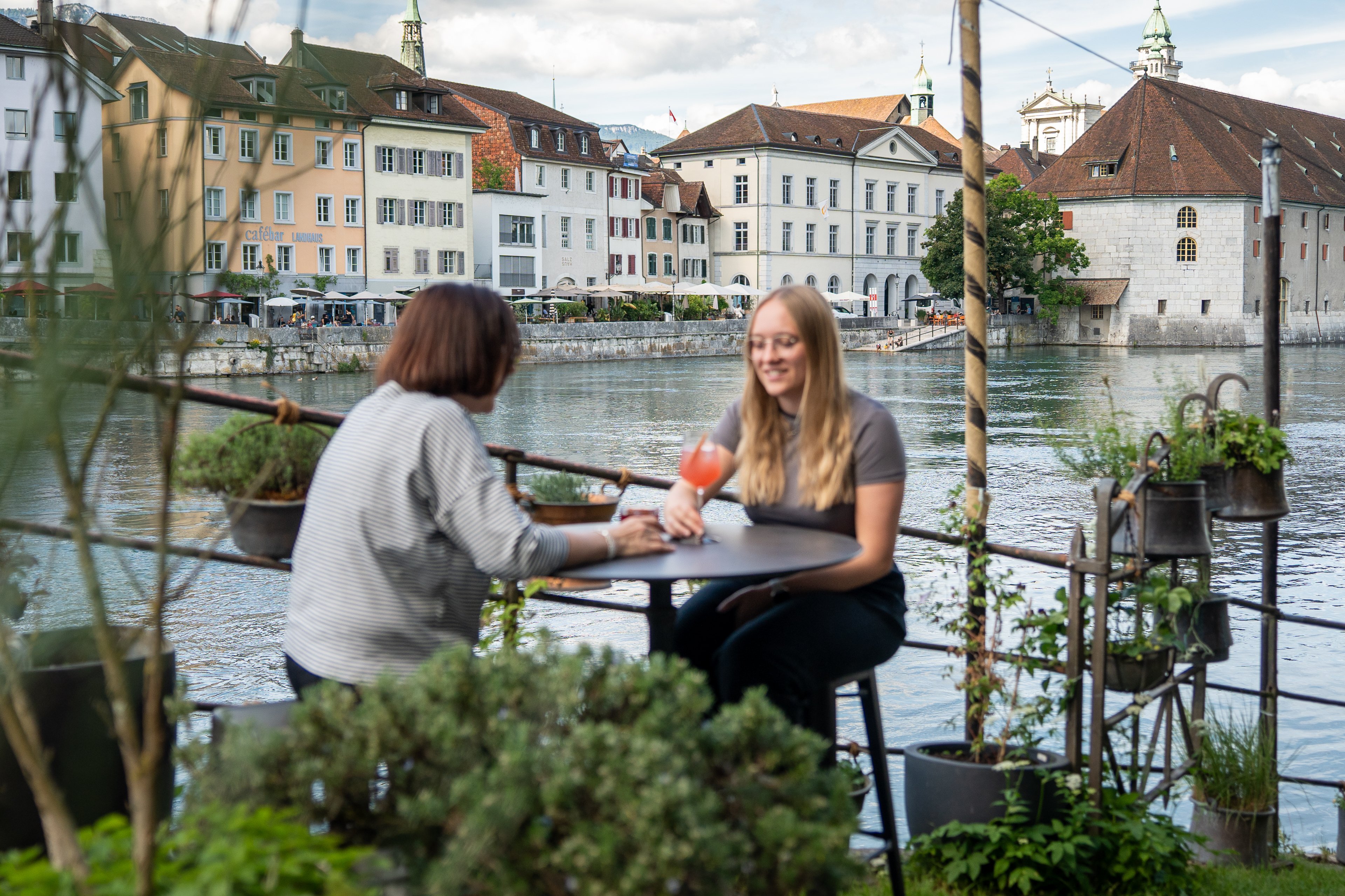 Zwei Personen, die je ein Getränk vor sich haben, an einem Tisch auf einer Terrasse an einem Fluss.