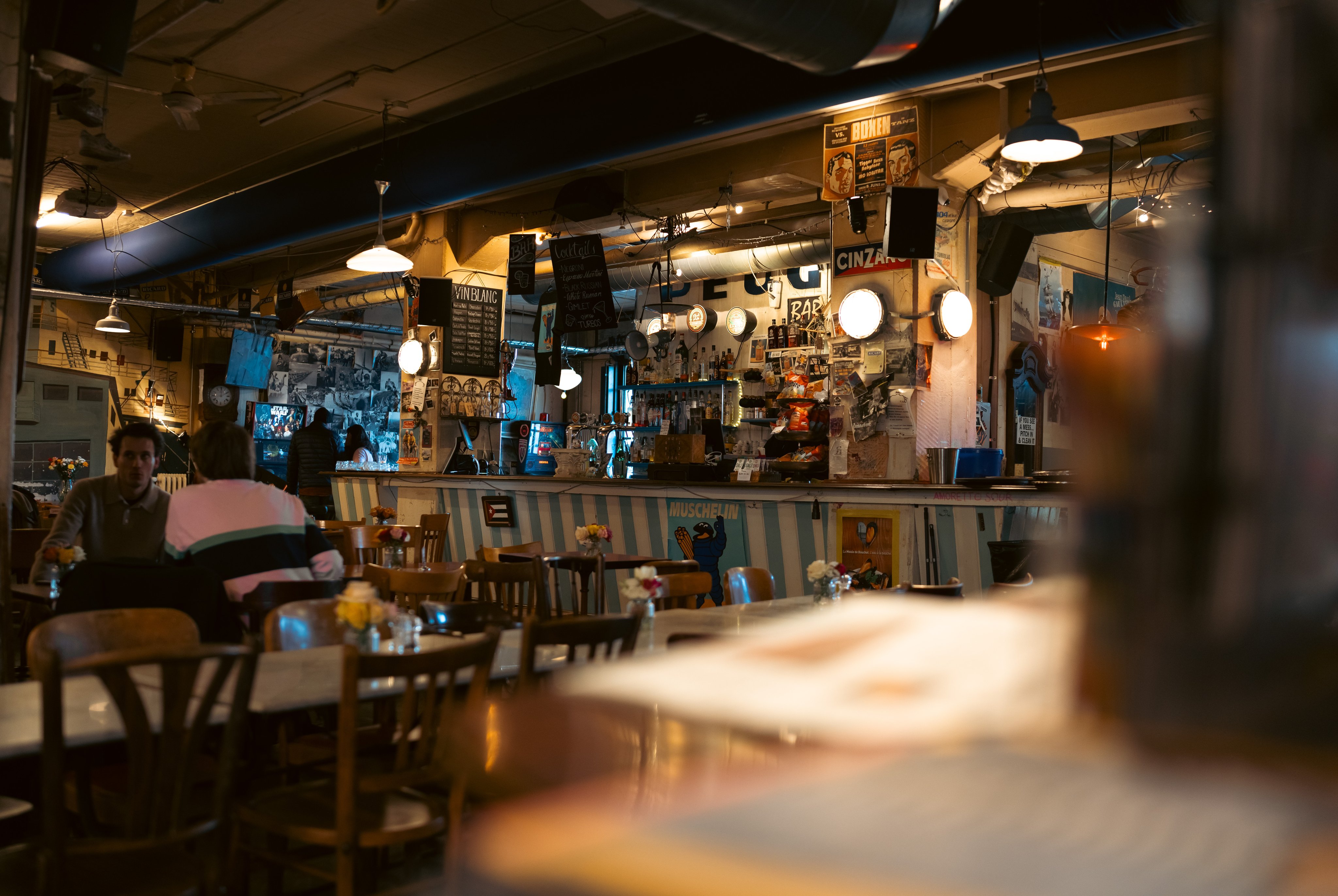 Comptoir de bar dans une salle de restaurant sombre. Les murs sont entièrement décorés d’affiches, de photos et de panneaux publicitaires. 