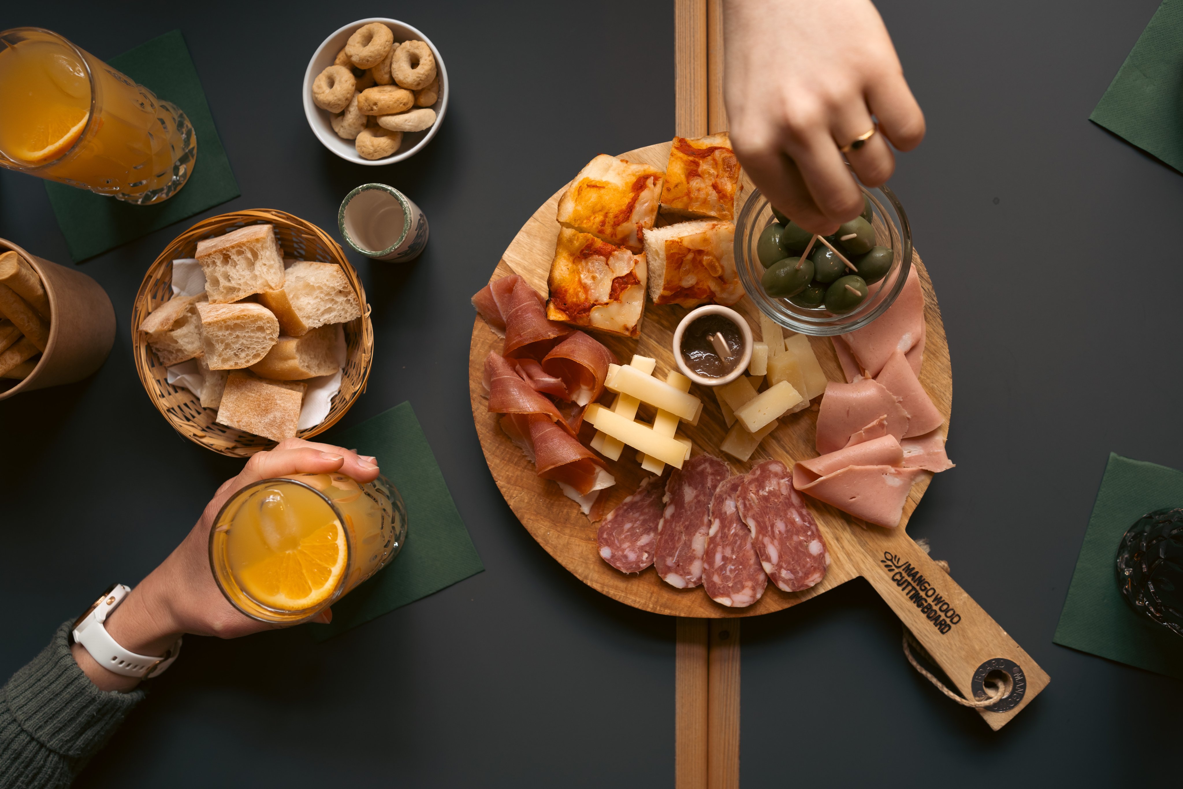 Un plateau posé sur une table avec assortiment de viandes, d’olives, de fromage et de pâtisseries. À côté: une corbeille de pain et des pâtisseries, ainsi que des boissons dans des verres.  