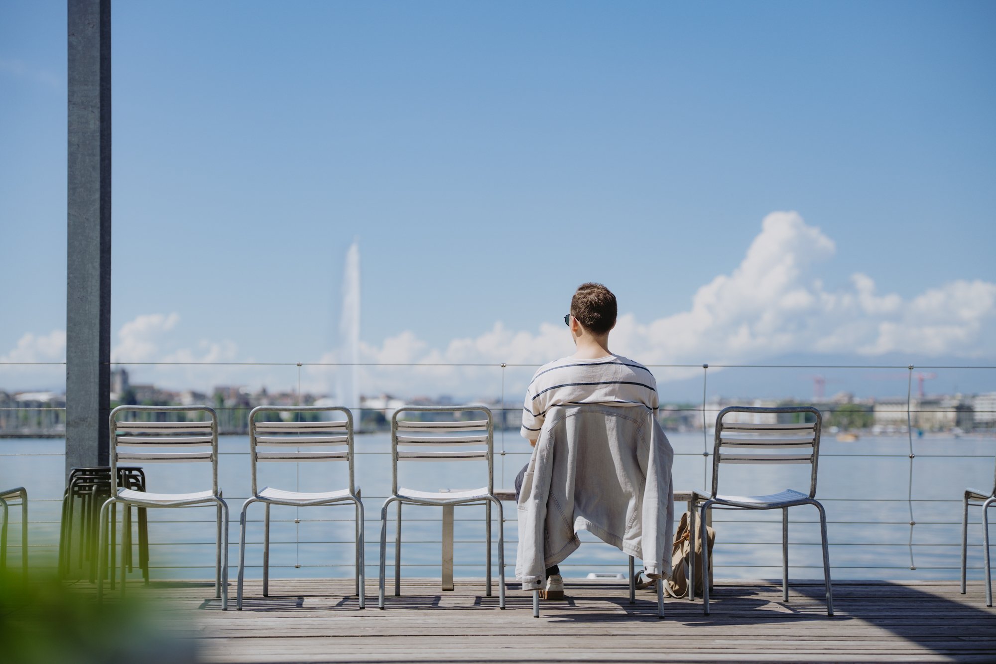 Une personne est assise sur une terrasse à côté de quatre chaises vides; à l’arrière-plan, on aperçoit le Léman et le jet d’Eau.