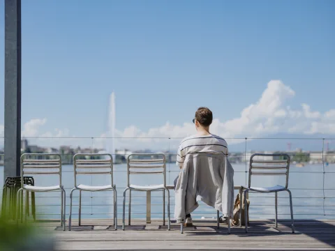 Une personne est assise sur une terrasse à côté de quatre chaises vides; à l’arrière-plan, on aperçoit le Léman et le jet d’Eau.