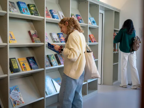 Eine Frau steht vor einem grossen Bücherregal und liest in einem Buch. Im Hintergrund steht eine zweite Frau.