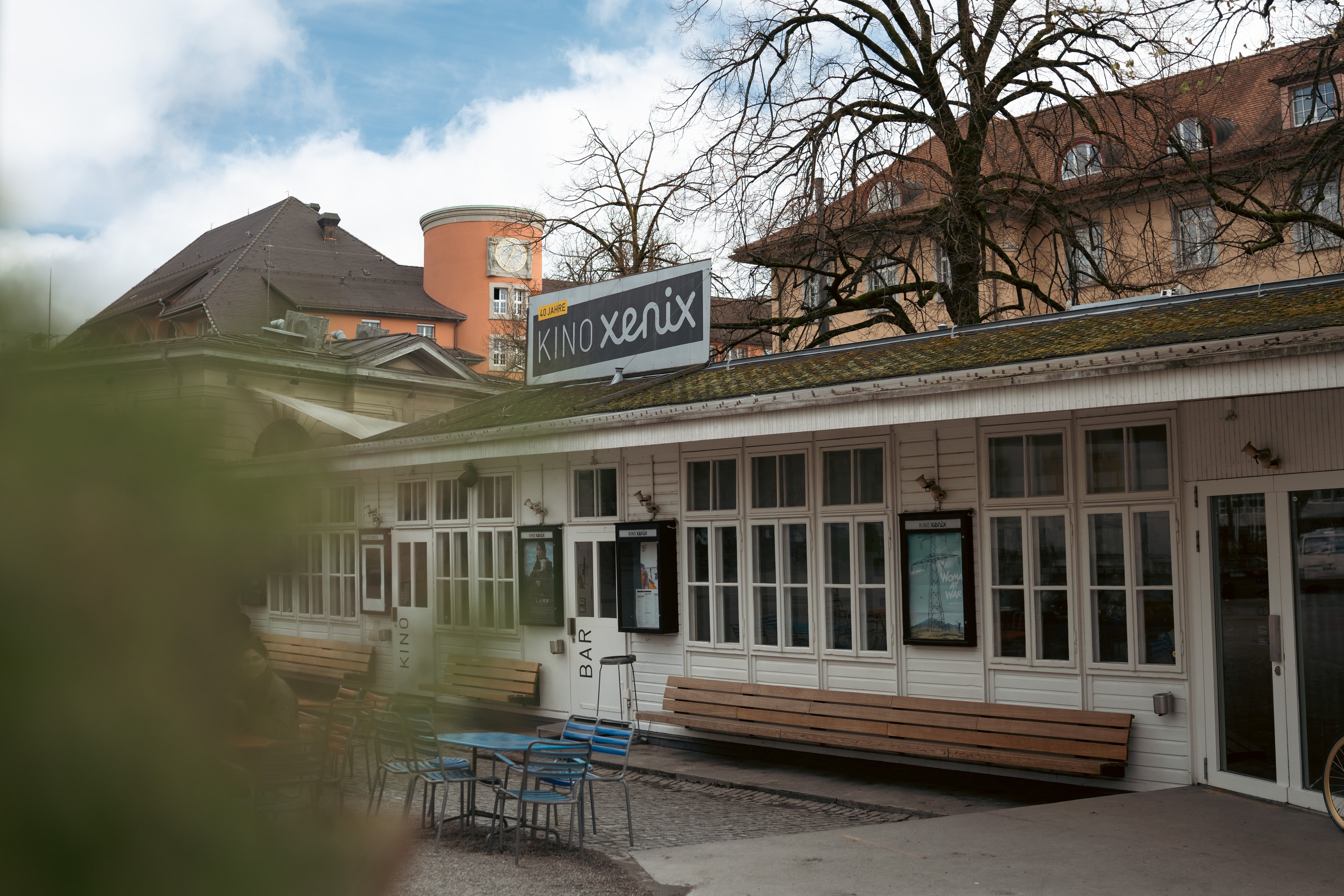 Un bâtiment plat et rustique en bois portant l’inscription cinéma et bar se trouve dans la cour intérieure. Des tables, chaises et bancs sont placés devant. 