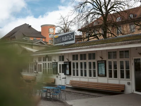 Un bâtiment plat et rustique en bois portant l’inscription cinéma et bar se trouve dans la cour intérieure. Des tables, chaises et bancs sont placés devant.