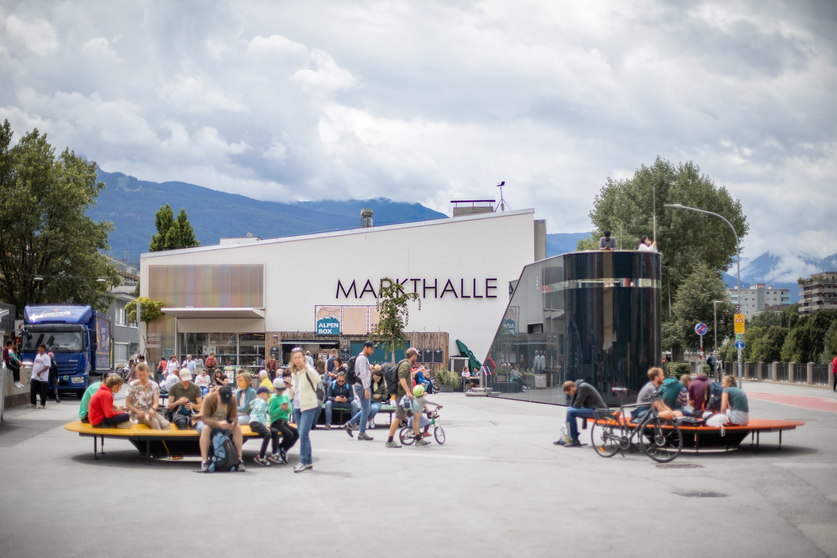 Menschengruppen sitzen auf Bänken auf dem Marktplatz vor dem Haus der Markthalle.