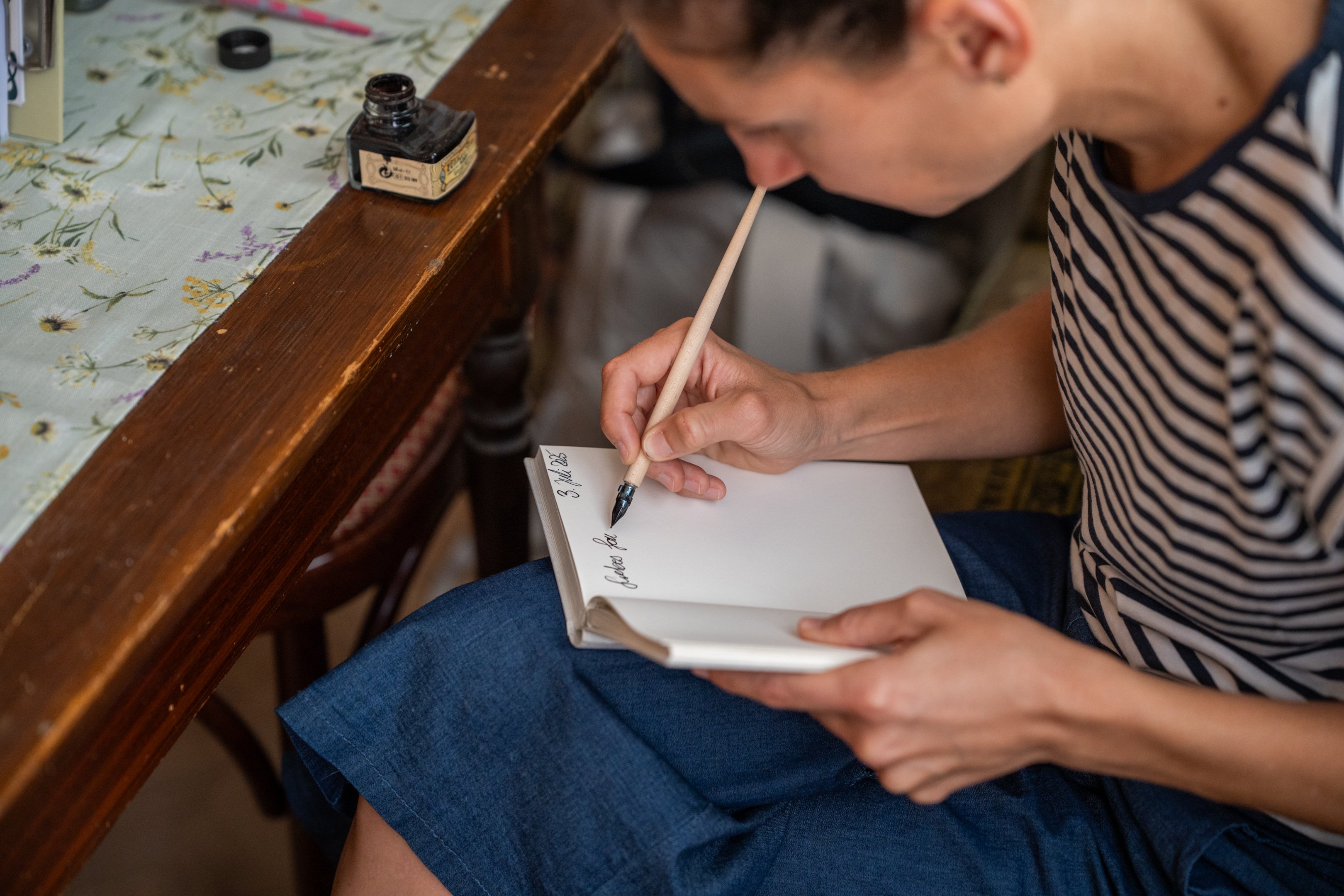 Une femme assise à une table et écrit dans un carnet blanc avec une plume.