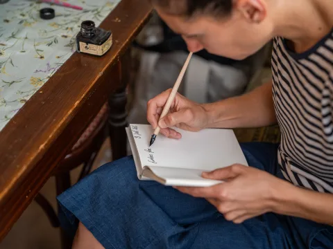 Une femme assise à une table et écrit dans un carnet blanc avec une plume.