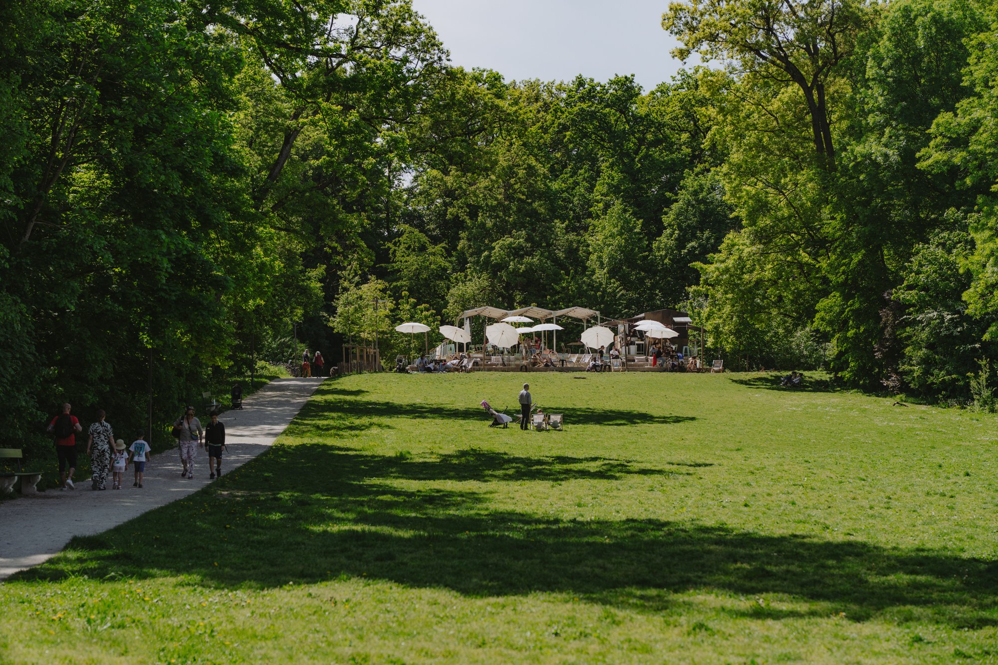 Une clairière avec des personnes dans la prairie et sur le chemin forestier; à l’arrière-plan, une buvette avec des parasols blancs.